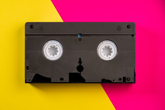 Black vintage VHS videotape cassette on yellow and pink background. Plastic retro video cassette with analog magnetic tape from 90s. Old technology for tape recording and watching media movies.