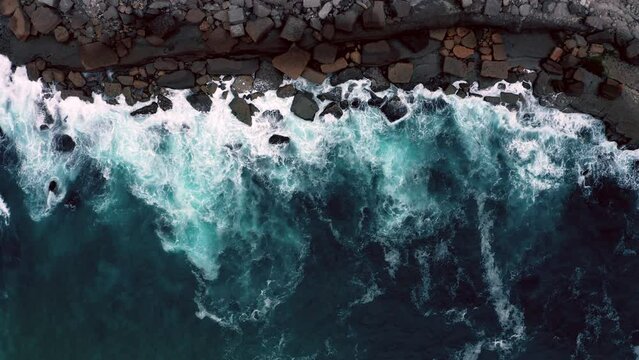 Aerial top view of the beautiful ocean rocky shore with rolling waves. Drone is hovering over rolling ocean waves to the rocky shore. Drone hovers over surface of the water along the rocky shore.