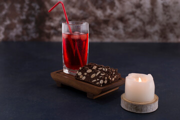 Red cocktail with ice cubes in a glass with a slice of cake on the wooden platter, side view