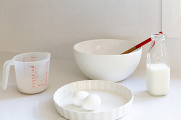High angle selective focus view of white cooking utensils and bowl, milk, eggs and flour in measuring cup set on quartz countertop