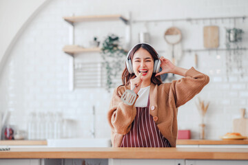 Happy carefree asian woman wearing headphones and ladle singing have fun moving listening to music relax in kitchen, She dance enjoy leisure weekend at home.