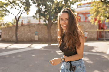 Excited lovely stylish woman with happy smile and loose wavy hair wearing black t-shirt and denim shorts walking around summer city in sunlight