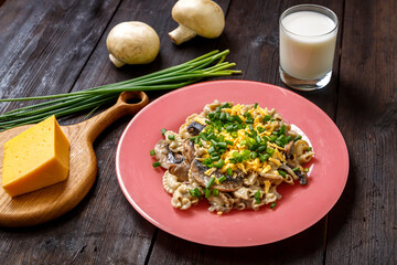 Pasta in a creamy sauce with mushrooms in a plate on a dark wooden table next to a glass of cream, cheese on the board, green onions and mushrooms.