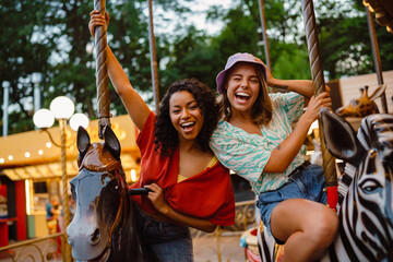 Young multiracial women riding on carousel in attraction park