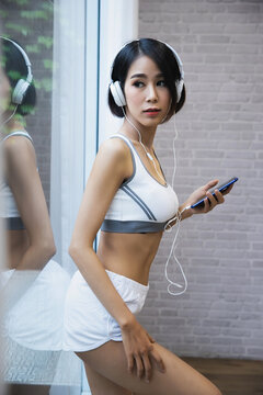 Asian woman in sportswear listening to music with headphone connected via smartphone after exercise at home fitness.