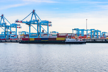 Cargo barge loaded with colourful containers sailing past a commercial dock with tall gantry cranes and stacks of containers
