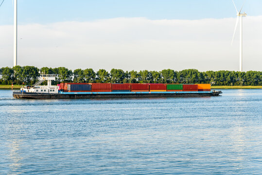 Cargo barge loaded with containers sailing up a river at sunset. Wind turbines are in background.