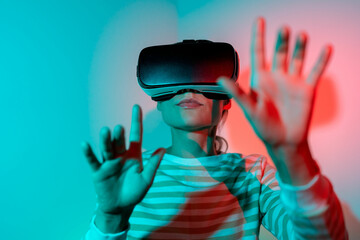 Young woman wearing VR glasses plays games with amazing graphics. Addicted to games girl illuminated by blue and red neon light. Female explores virtual reality using goggles. 