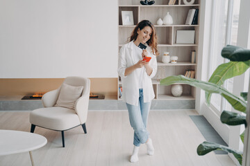 Female holds bank credit card, phone makes cashless payment for house utilities in modern apartment