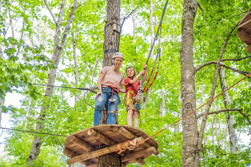 Naklejka na ściany i meble Mother and son climbing in extreme road trolley zipline in forest on carabiner safety link on tree to tree top rope adventure park. Family weekend children kids activities concept