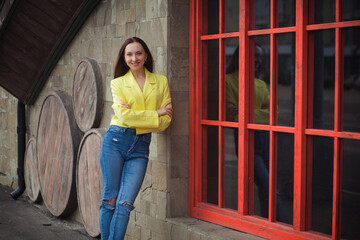 A stylish slim smiling woman stands near a building with wooden red windows and a stone wall, on a street in the city. She is wearing blue jeans and a short yellow leather jacket.