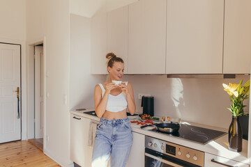 Smiling young woman holding cup of coffee near breakfast in kitchen.