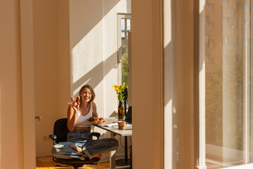 Smiling woman holding croissant and looking at camera near laptop and flowers at home.