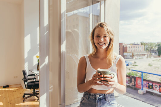 Happy blonde woman in top holding cup and looking at camera near open window at home.
