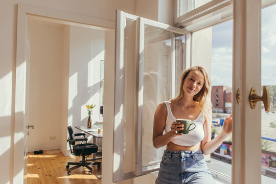 Smiling woman in top holding cup near open window at home.