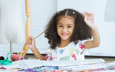 Cute African little girl drawing or sketching clothing design on paper at tailor room, cozy home as...