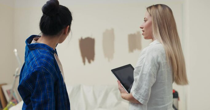 Two women from the back, pointing with their fingers at a room undergoing renovation. The room has a sofa, a ladder, boxes, paintings, brushes and other tools.