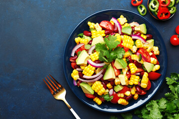 Mexican spicy salad with corn, red beans, avocado, jalapeno peppers, cherry tomatoes, onion and cilantro. Blue table background, top view