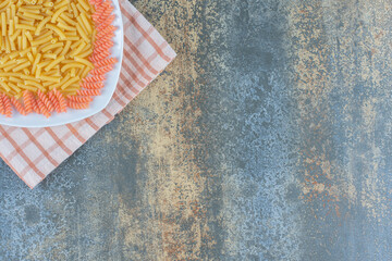 Uncooked penne and fusilli pasta in bowl on towel, on the marble background