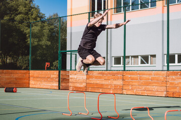 Blond boy in sportswear jumps over red obstacles to improve lower body dynamics. Plyometric training in an outdoor environment. Improve your skills