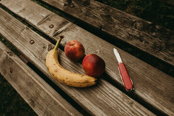 Two peaches and a banana forming a smile on a wooden bench, with a pocket knife. - obrazy, fototapety, plakaty