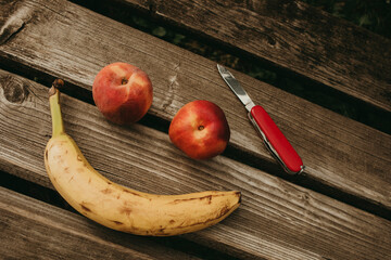 Two peaches and a banana forming a smile on a wooden bench, with a pocket knife. - obrazy, fototapety, plakaty