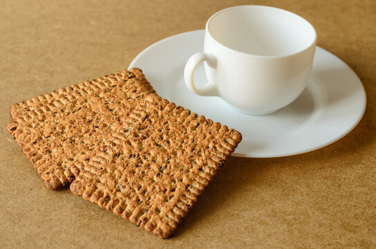 Three crackers with roasted flax seeds, leaning on a white saucer with a white cup standing on it, surface of chipboard table, selective focus, close-up