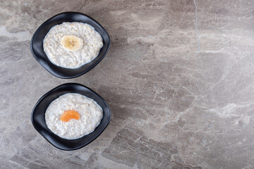 Sliced oranges on a bowl of porridge next to sliced banana on a bowl of porridge , on the marble background