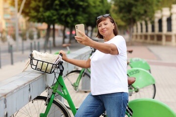 Fototapeta na wymiar Weekend ride on bicycle. Mature woman renting bike, using smartphone for selfie. Concept of online payment, commute in the city and shared transport. Travel, tourism in summer. Eco friendly transport