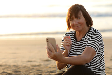 Fototapeta na wymiar Smiling mature caucasian woman listening music in headphones looking at camera relaxing and resting on sandy beach by the sea at sunrise. Relaxation concept