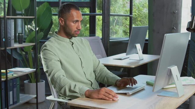 Shot of young attractive focused smart African man sitting at workplace in modern office, typing on keyboard of computer, using mouse. Office worker showing approval gesture to camera. Thumbs up