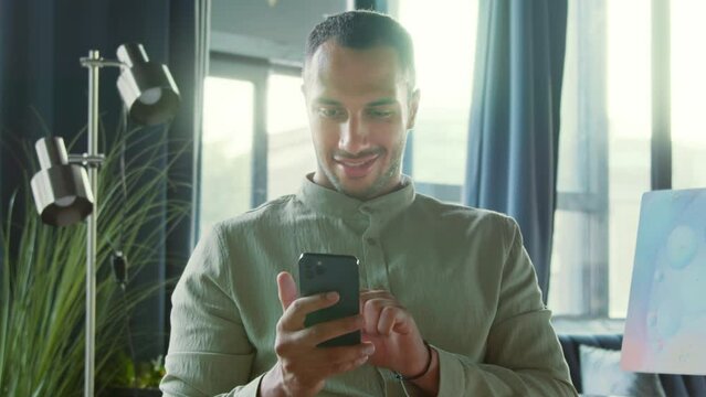 Portrait of pleasant-looking happy young man in mint outfit holding smartphone, looking at screen, scrolling. Footage of smiling guy chatting, using gadget at work. Office, good news. Indoors