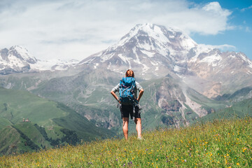Backpacker woman with a backpack standing and enjoying snowy slopes of Kazbek 5054m mountain while she walking by green grass hill. East Caucasus mountains, Georgia.