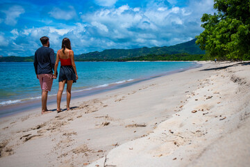 a couple in love holding hands walk along a paradise beach with turquoise water, white sand and palm trees in the background; romantic walk on a wonderful tropical beach in costa rica
