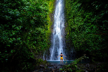 brave girl stands in front of a mighty waterfall with her hands raised in the air; celebration of a successful climb over a waterfall; tropical waterfall in Costa Rica; hidden gems of costa rica