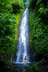 brave girl stands in front of a mighty waterfall with her hands raised in the air; celebration of a successful climb over a waterfall; tropical waterfall in Costa Rica; hidden gems of costa rica