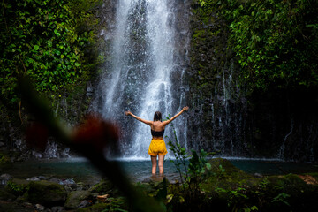 brave girl stands in front of a mighty waterfall with her hands raised in the air; celebration of a...
