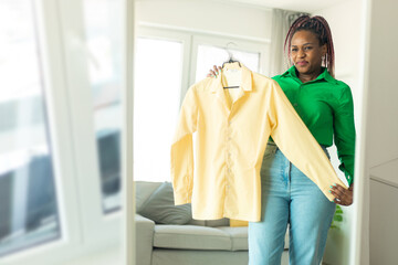 African american lady choosing clothes to wear, looking herself in mirror in living room at home, free space