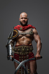 Shot of muscular gladiator from past dressed in armor and red cape against gray background.