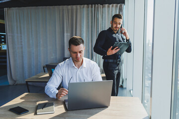 Two male focused colleagues discussing business project looking at laptop screen during meeting in modern meeting room with glass walls