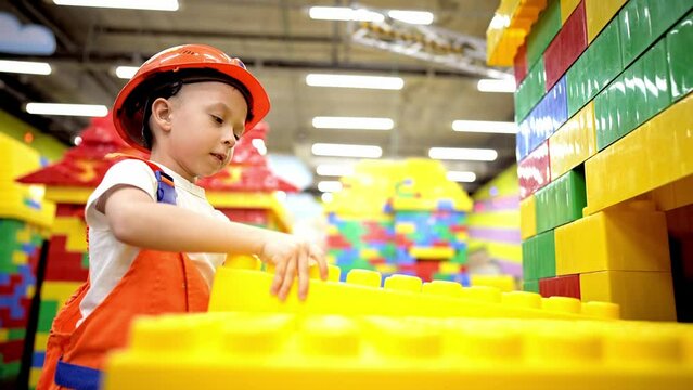 Baby boy stands in helmet and clothes of builder and makes house out of constructor bricks. Game in profession in children. Construction of toy building. Choosing future profession in childhood.