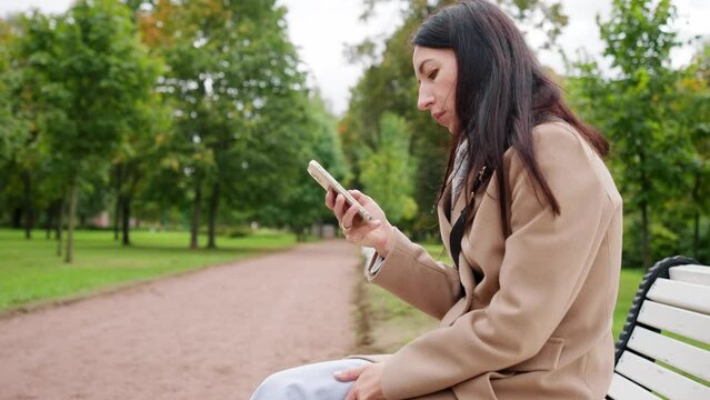 A young woman sits on a bench with a phone in her hands. In the background green park.