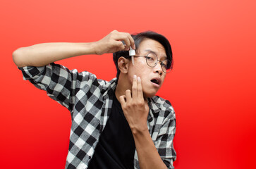 asian young man applying beauty acne serum and smiling at camera isolated over red background