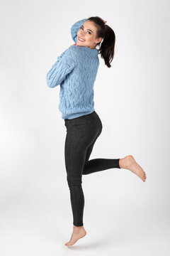 Fashion and lifestyle concept. Studio portrait of beautiful woman wearing black jeans and blue sweater in white studio background. Model jumping in air with bare feet. Image contains motion blur