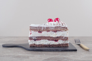 Small cake slice with cream, cherry and cocoa powder topping on a board on marble background