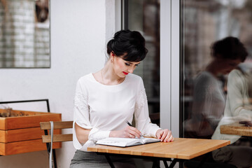 Serious focused smart businesswoman is writing notes on notepad, making agenda on personal organizer. Female in business suit sitting at table at cafe on terrace, work and event planning.