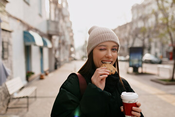 Funny adorable girl in knitted cap and black jacket winking at camera and bitting cookie and...