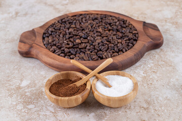 Ground coffee and a sugar bowls next to coffee beans piled on a wooden tray on marble background