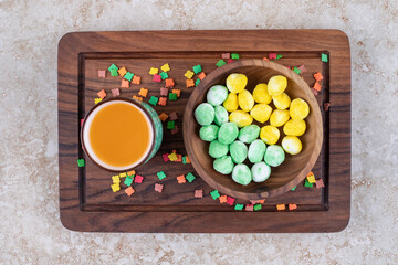 A bowl of mint and lemon candies and a cup of juice on a wooden board on marble background