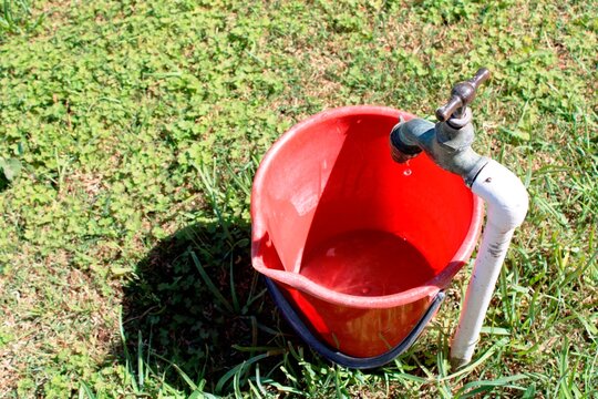Cubo rojo bajo el viejo grifo donde el agua sale en gotas debido a la escasez de la sequ&iacute;a, forma un hermoso y original dise&ntilde;o natural con un fondo de hierba verde